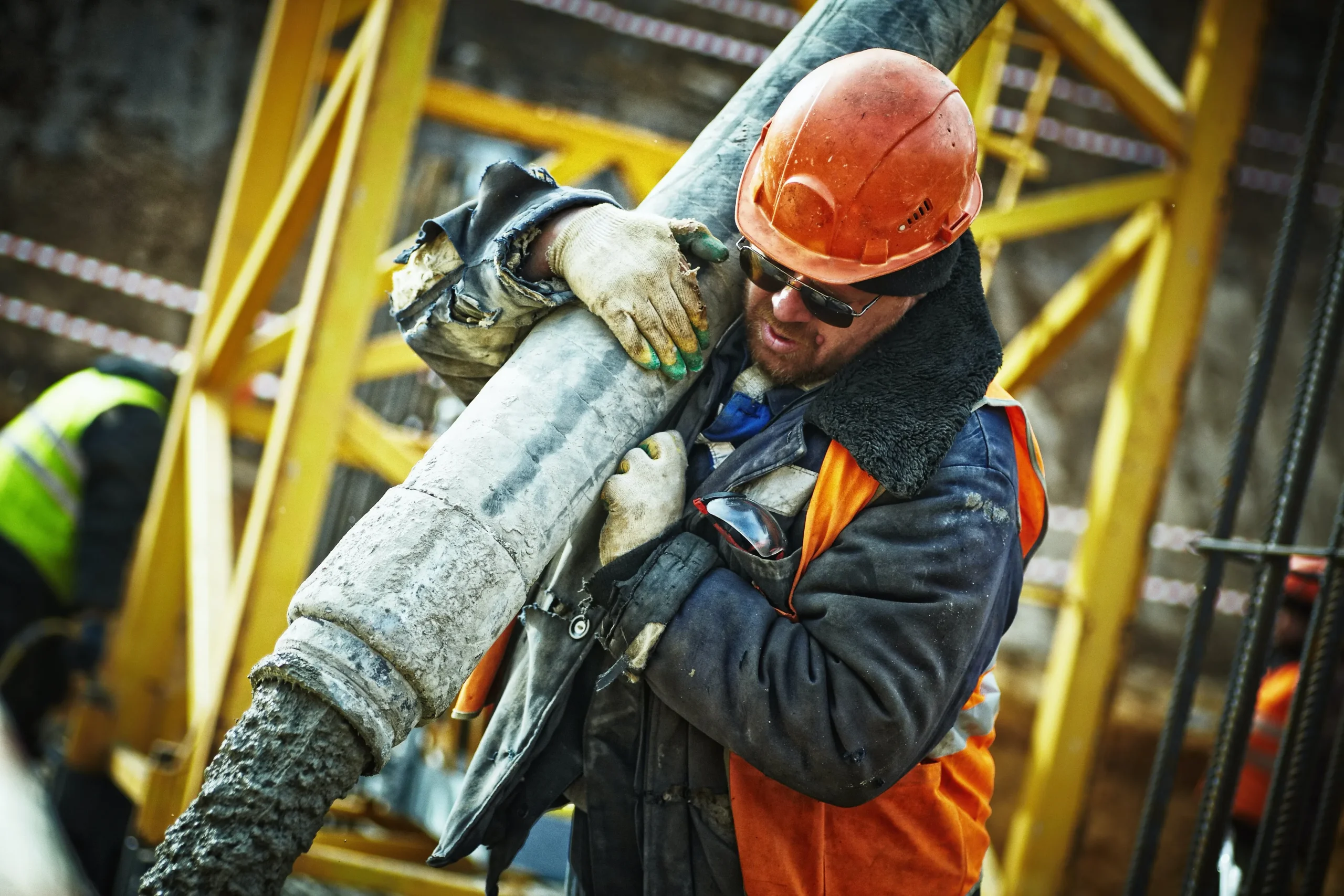 Truck driver wearing a thick coat, gloves, a hard hat, and an orange hazard vest holds a tube on his shoulder directs wet concrete into a form on the ground after completing the DOT SAP program in Texas.